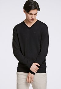 Schwarzer V-Ausschnitt-Pullover mit Strickstruktur, ausgestattet mit langen Ärmeln und einem taillierten Saum. Dezentes Logodetail in der Nähe des Halsausschnitts. Kombiniert mit hellbeigen Hosen.