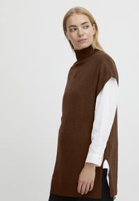 Brun turtleneck ærmeløs sweater lagdelt over en hvid langærmet skjorte, med sideslids og en blød tekstur.