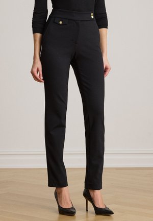 Femme portant un pantalon noir ajusté avec des boutons dorés à la taille, associé à des chaussures noires à talons hauts, debout sur un sol en bois.