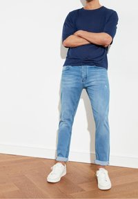 Jean en denim bleu clair avec légère usure, associé à un t-shirt bleu marine et des baskets blanches. Ourlets des jeans retroussés.