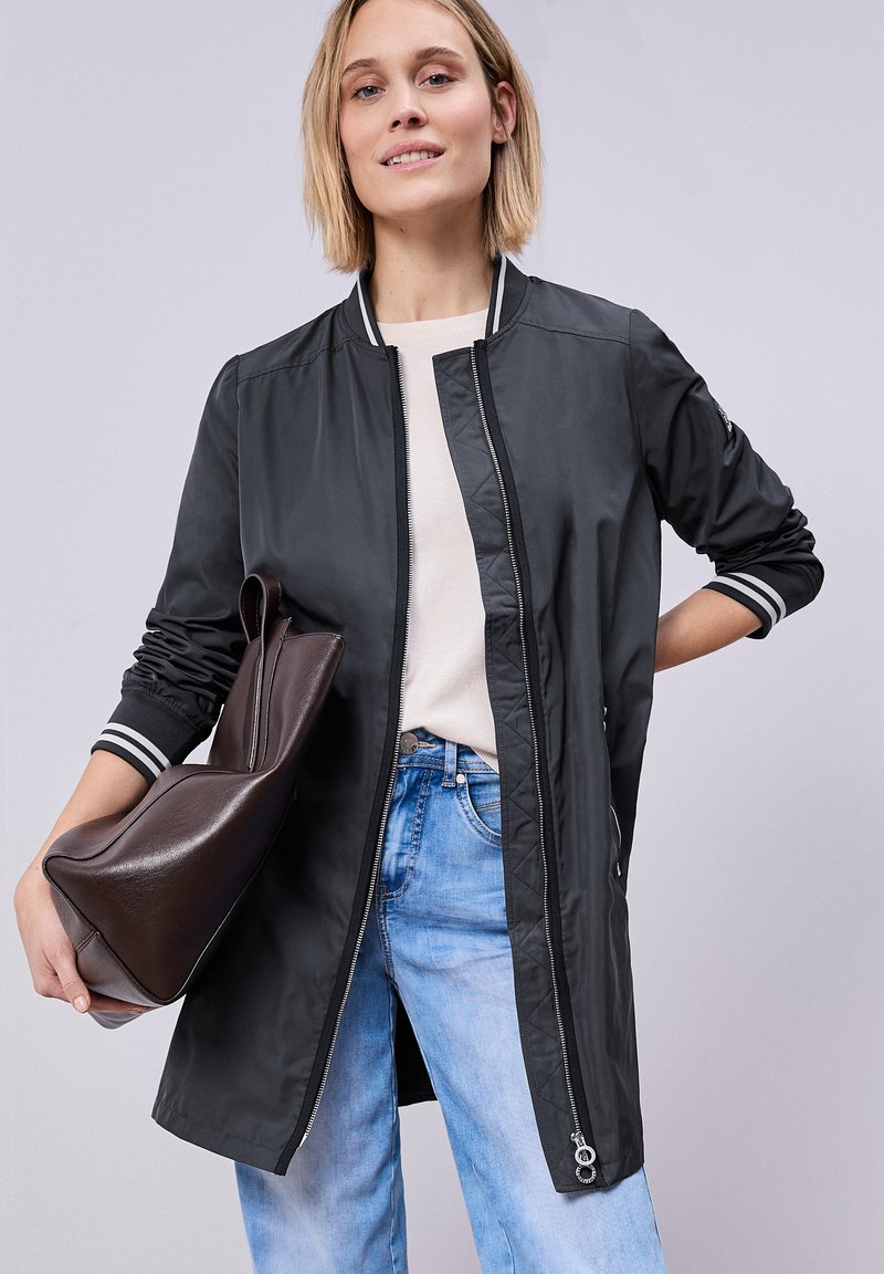 Mujer con una chaqueta bomber negra larga sobre una camiseta blanca y vaqueros azules, sosteniendo un bolso grande de cuero marrón oscuro en una mano.