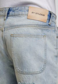 Šviesiai mėlyni džinsiniai šortai su nusidėvėjusiu kraštu, turintys galinį kišenę ir šviesiai rudos odos ženklelį su įspaustu užrašu "2Y STUDIOS." Siuvimo linijos kontrastingos spalvos.