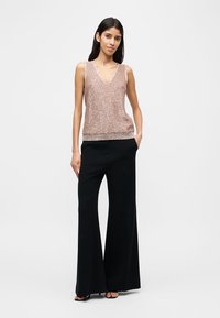 Γυναίκα που στέκεται φορώντας ρόζ μπλούζα με παγέτες χωρίς μανίκια και μαύρο παντελόνι με φαρδιά πόδια και ψηλή μέση, με σανδάλια, τα χέρια στις τσέπες.