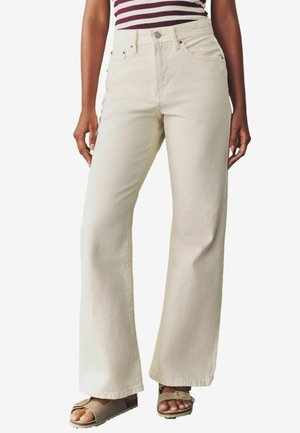 Femme portant un pantalon crème à taille haute et jambes larges avec poches avant et fermeture à boutons, associé à des sandales beiges à bout ouvert.