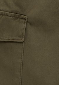 Gros plan sur un tissu vert olive avec une poche à rabat rectangulaire cousue sur le côté gauche, montrant la texture détaillée du textile et les lignes de couture.