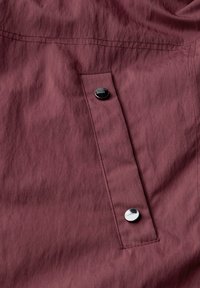 Burgundy-kankaan pehmeä tekstuuri on varustettu läpällisellä taskulla, jota korostavat kaksi hopeista nepparipainiketta ja näkyvät ompeluvarat.
