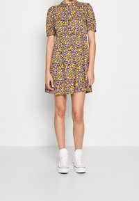 Blommig klänning med puffärmar, rund halsringning och utställd kjol. Färgerna inkluderar gult, lila och svart på en ljus bakgrund.