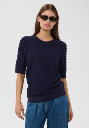 Vrouw met lang bruin haar, zwarte zonnebril, een gestructureerde marineblauwe korte mouw trui en blauwe jeans, staand tegen een effen achtergrond.