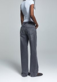 Mujer vestida con jeans gris claro, camiseta blanca de manga corta ajustada y zapatos planos oscuros, de pie con las manos en los bolsillos contra un fondo gris.