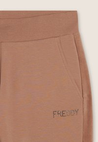 Pantalones de chándal color marrón claro con una textura suave, que cuentan con bolsillos laterales y un logo "FREDDY" decorado con cristales en la parte delantera izquierda.