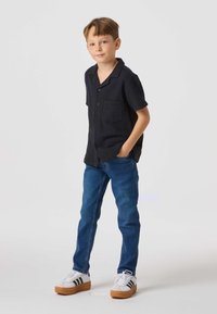 Sort kortærmet skjorte med knapper og lomme, blå jeans og hvide sneakers med sorte striber og beige såler. Ensfarvet hvid baggrund.