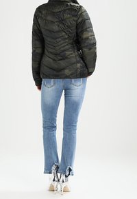 Veste camouflage rembourrée avec une silhouette cintrée et un matelassage diagonal, assortie à un jean bleu clair avec des ourlets effilochés et des talons hauts argentés.