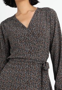 Robe portefeuille à imprimé léopard en gris avec des taches noires et orange. Dispose de manches longues, d'un décolleté en V et d'une ceinture nouée à la taille. Texture de tissu douce.