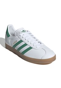 White leather sneakers feature three green stripes and a gum rubber sole. A green logo is visible on the tongue, with "GAZELLE" embossed.