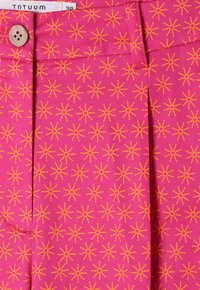 Klarrosa tyg med små upprepade orange solstrålmönster, träknapp och vit kläddetikett som visar storlek 38.