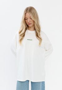 Witte oversized t-shirt van zachte katoen met groene tekst "MIRONS" gecentreerd op de borst. Lange mouwen en een ontspannen pasvorm design.