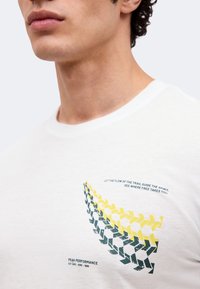 Κοντινό πλάνο ενός άνδρα που φοράει λευκό πουκάμισο με κίτρινα και σκούρα πράσινα αφηρημένα σχέδια βελών και κείμενο κοντά στην περιοχή του στήθους.