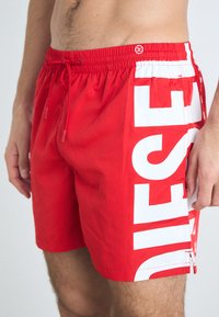 Bermuda de bain rouge en tissu léger avec un imprimé logo blanc sur le côté et une taille élastique avec cordon de serrage.