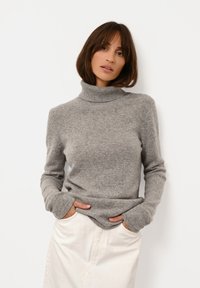 Grå halsringad tröja i ett mjukt, texturerat material. Har långa ärmar och en avslappnad passform, bärs över en ljusfärgad kjol.