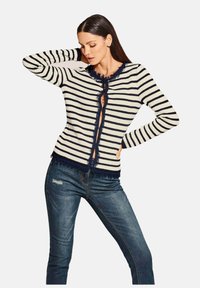 Gestreepte cardigan in crème en marineblauw, met een franjeszoom, ronde halslijn en knoopsluiting. Gecombineerd met slim-fit donkere spijkerbroek.