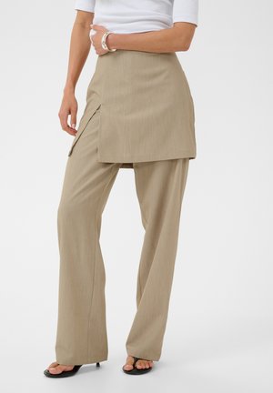 Donna che indossa pantaloni beige a gamba larga con sovrapposizione di gonna a portafoglio attaccata e tacchi aperti neri, in piedi davanti a uno sfondo uniforme.