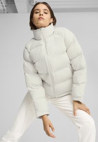 Světle šedá puffer bunda s vysokým límcem, zipovým zapínáním a prošívaným designem; nošena přes bílé kalhoty, s texturou, která vypadá měkce a polstrovaně.