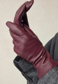 Burgundy leather glove with a fitted design and gathered wrist, displayed against a gray textured fabric background.