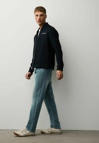 Jeune homme marchant, portant une veste noire zippée, un jean bleu clair et des baskets beiges, sur un fond gris clair uni.