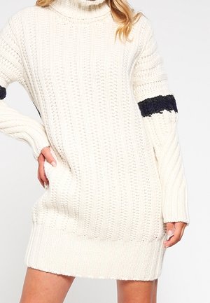 Kobieta ubrana w biały dziany sweter z golfem, o długich rękawach, z czarnym paskiem na lewym ramieniu.