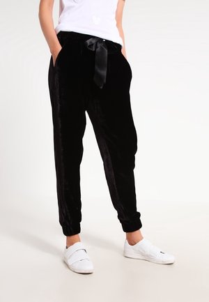Person iført sorte fløjls joggerbukser med satin snøre og hvide slip-on sneakers, stående med hænderne i lommerne.