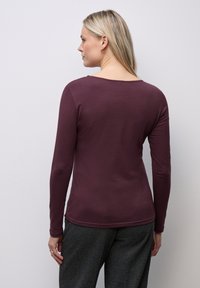 Maglietta a maniche lunghe di color bordeaux realizzata in un materiale morbido. Presenta un ampio scollo e un design aderente, abbinata a pantaloni grigi con texture.