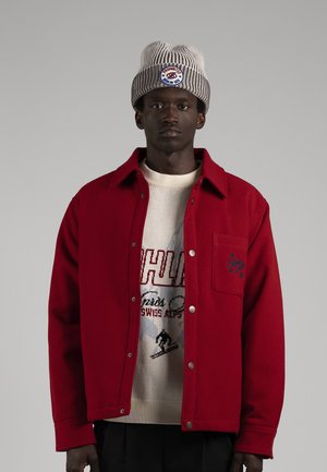 Un homme pose avec une veste rouge boutonnée, un pull crème à motif graphique, un pantalon noir et un bonnet en maille rayé, sur un fond gris uni.