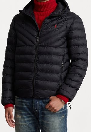 Mand iført en sort quiltet dunjakke med hætte, rød kabelstrikket rullekravesweater og slidte blå jeans.