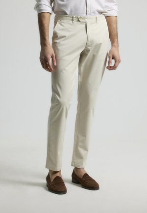 Pantalones beige de corte slim con una textura suave, con cierre de botón, trabillas para el cinturón y bolsillos, combinados con mocasines marrones.