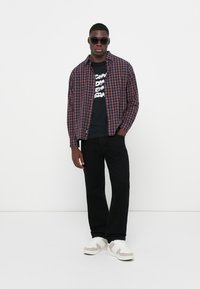 Checkered button-up shirt in navy and red over a black graphic t-shirt, paired with black pants and white sneakers. Model wears sunglasses.