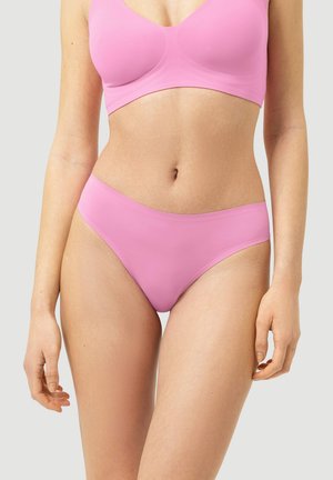 Femme portant un soutien-gorge et des sous-vêtements roses assortis sans couture, debout devant un fond uni clair.