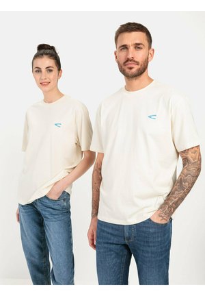 T-shirts en coton couleur crème à manches courtes, ornés d'accents de logo bleu clair, portés avec un jean bleu par deux mannequins.