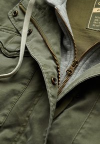 Olivgrön jacka med grå hoodie-inredning; har en dragkedja fram med gyllene dragkedja och snodddekorationer, texturerad tyg.