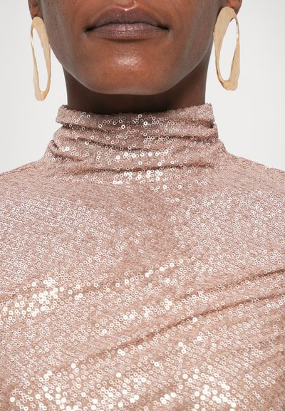 Ροζ χρυσό τοπ με παγιέτες και ψηλό λαιμό, διαθέτοντας μαλακή υφή και λαμπερή επιφάνεια, συμπληρωμένο με μεγάλα αφηρημένα χρυσά σκουλαρίκια.