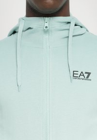 Lyseblå hettegenser med glidelås laget av myk stoff, med glidelås, snorer og svart EA7 Emporio Armani-logo på fronten.