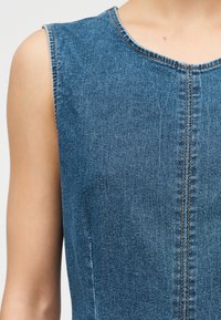 Αμάνικο τοπ από τζιν με ορατές πορτοκαλί ραφές κατά μήκος της μασχάλης, του λαιμού και της κεντρικής ραφής σε βραχίονα και ώμο με ανοιχτό τόνο δέρματος.