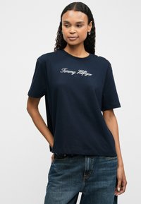 Tricou din bumbac bleumarin cu mâneci scurte, având brodat "Tommy Hilfiger" în alb și accente de buton aurii pe umăr.
