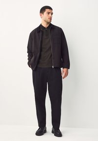 Zwarte jas met ritssluiting, daaronder een getextureerd donker overhemd en zwarte broek. Glad materiaal, eenvoudig ontwerp en minimale hardware-details.