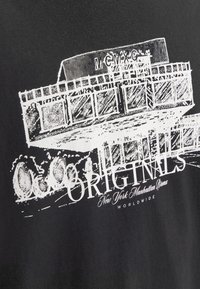 Camiseta negra con diseño gráfico blanco que presenta un edificio y el texto "ORIGINALS Nueva York Manhattan Bronx". Estilo vintage, letras en negrita.