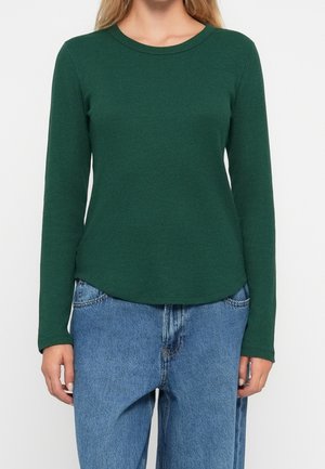 Kobieta ubrana w dopasowaną, ciemnozieloną, prążkowaną bluzkę z długim rękawem, w połączeniu z luźnymi niebieskimi dżinsami, stojąca na jednolitym białym tle.