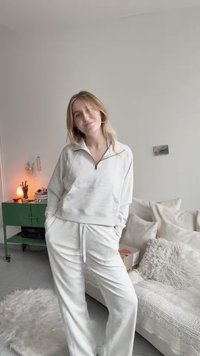 Jasnoszara, zapinana na zamek bluza z kieszenią z przodu oraz pasujące dresy. Miękka, teksturowana tkanina w luźnym kroju. Pomieszczenie o neutralnej dekoracji.