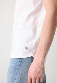 Camiseta de algodón blanca con cuello redondo y mangas cortas, que presenta un parche con logo en rojo y beige cerca del dobladillo. Combinada con jeans de mezclilla azul.
