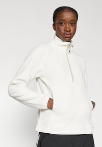 Λευκό φλις πουλόβερ με μισό φερμουάρ, ραγκαλένια μανίκια και μπροστινή τσέπη. Μαλακή υφή, χαλαρή εφαρμογή. Συνδυασμένο με μαύρο παντελόνι.