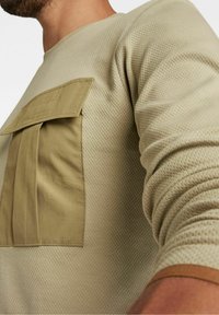 Beige strukturiertes Hemd mit einer großen, umgeschlagenen Tasche in Khaki auf der linken Brust. Gerippte Bündchen und Halsausschnitt mit vertikalem Muster.