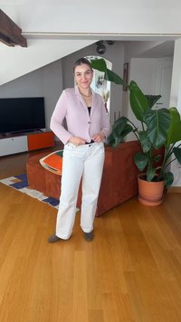 Γυναίκα που χαμογελά και στέκεται στο σαλόνι, φοράει ανοιχτό ροζ σακάκι, άσπρο παντελόνι, με μεγάλο πράσινο φυτό και πορτοκαλί καναπέ πίσω της.